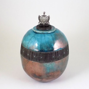 May include: A round ceramic urn with a teal and copper glaze. The urn has a dark brown band with decorative accents and a silver metal top with a winged design.