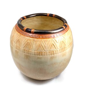 May include: A beige ceramic vase with a rounded shape and a wide opening. The rim is decorated with a dark band and orange and black accents. The body features carved geometric patterns, creating a textured surface.