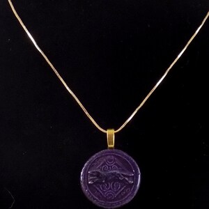 May include: A gold-toned chain necklace featuring a circular, deep purple pendant. The pendant has a raised, embossed design of a leaping animal figure centered within a geometric pattern, suspended from a simple gold-toned metal bail.