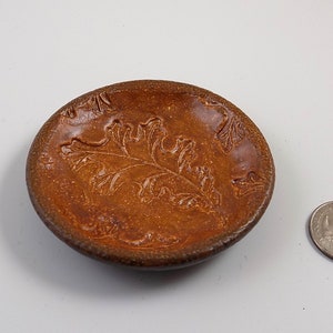 Può includere: Un piccolo piatto rotondo in ceramica marrone con un motivo a foglia in rilievo. Il piatto è circa delle dimensioni di una moneta da 20 centesimi.