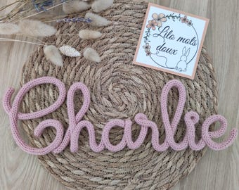 May include: A pink, crocheted name, "Charlie", is displayed on a woven, circular placemat. A small card with floral illustrations and the text "Lilo mots doux" is visible. Dried flowers and decorative elements are arranged around the name.