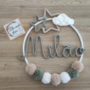 May include: A handcrafted nursery decoration with a white circular frame, featuring the name "Milão" in grey yarn. The design includes a star, cloud, and pom-poms in neutral tones. A small card with text and a bunny illustration is also present.