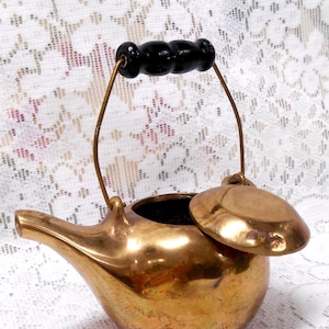 May include: A small, vintage brass teapot with a black wooden handle and a hinged lid. The teapot has a curved spout and a rounded body. The handle is attached to the body with brass wire. The lid is slightly ajar, revealing the interior.