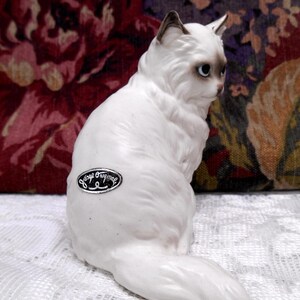 May include: White porcelain figurine of a sitting cat with a fluffy tail. The cat has blue eyes and a black nose. The figurine is marked "Vintage Original" on the bottom.