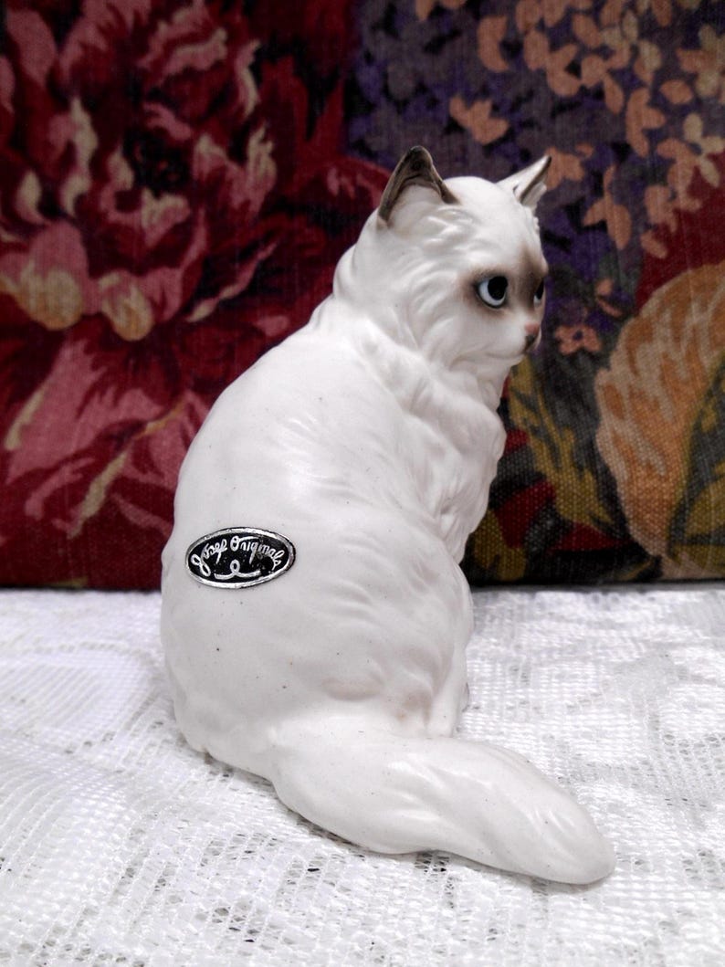 May include: White porcelain figurine of a sitting cat with a fluffy tail. The cat has blue eyes and a black nose. The figurine is marked "Vintage Original" on the bottom.