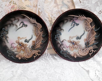 May include: Two decorative plates with a black background and gold dragon designs. The dragons are detailed with blue eyes and are flying in a swirling cloud pattern.