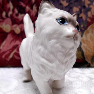 May include: White porcelain figurine of a cat with blue eyes. The cat is standing on all four paws and has a fluffy tail.