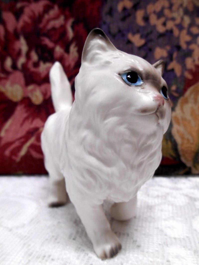 May include: White porcelain figurine of a cat with blue eyes. The cat is standing on all four paws and has a fluffy tail.