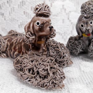 May include: Two ceramic poodle figurines. One is lying down, the other is sitting. Both have brown and gray fur, with a textured, curly appearance. They have black eyes and small yellow collars. The background is a white, lacy fabric.