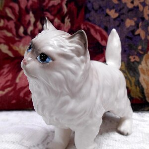 May include: White porcelain figurine of a cat with blue eyes. The cat is standing on all four paws and has a fluffy tail.