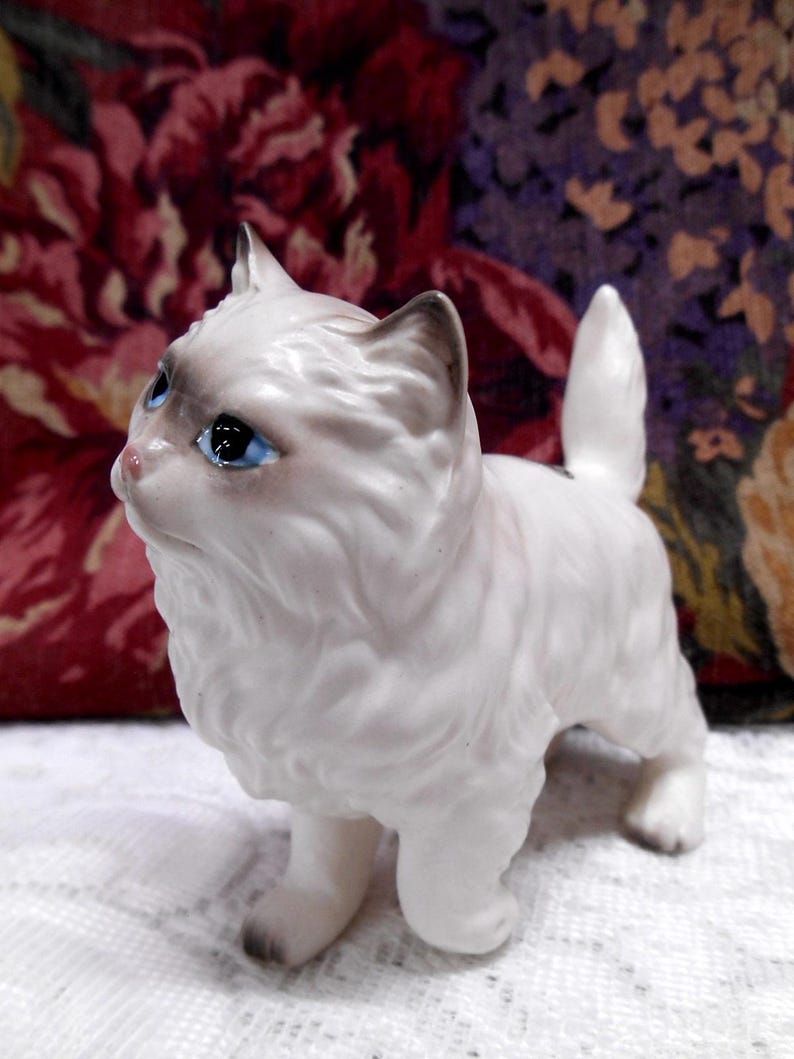 May include: White porcelain figurine of a cat with blue eyes. The cat is standing on all four paws and has a fluffy tail.