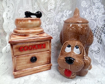 May include: Two ceramic cookie jars. One is a light brown, rectangular jar with the word "COOKIES" in red. The other is a brown dog-shaped jar with a lid. The dog has a red tongue and is looking up.