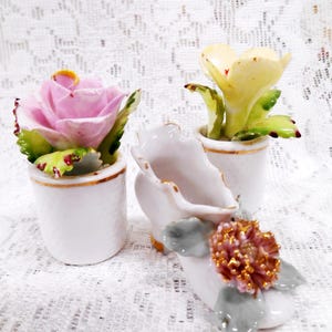 May include: A collection of delicate porcelain flower figurines. The set includes a pink rose, a yellow flower, and a pink and gold flower arrangement, all in small white pots with gold trim. The flowers have green leaves.
