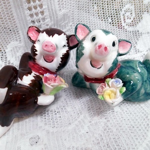 May include: Two ceramic pig figurines, one brown and white, the other green and white, each holding a bouquet of flowers. Both pigs are lying down and wearing red scarves. The figurines have pink ears and snouts.