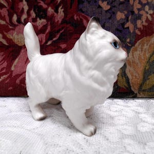 May include: A white porcelain figurine of a cat with blue eyes. The cat is standing on all fours and has a fluffy tail.