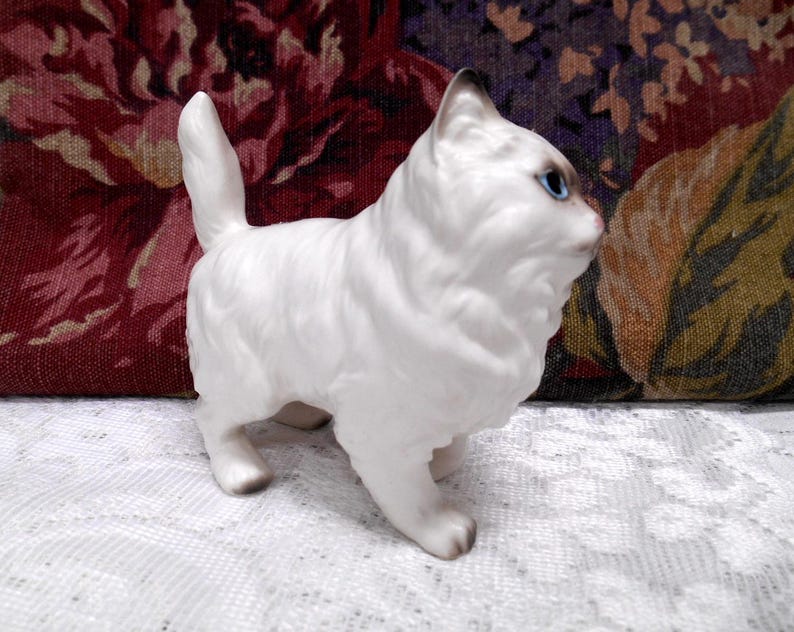 May include: A white porcelain figurine of a cat with blue eyes. The cat is standing on all fours and has a fluffy tail.