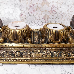 May include: Antique brass double inkwell with ornate detailing. The inkwells have hinged lids and white ceramic ink pots. The base features intricate floral and scrollwork designs, showcasing a vintage aesthetic.