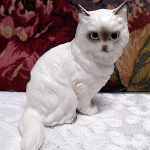May include: White porcelain figurine of a sitting cat with grey eyes and a fluffy tail.