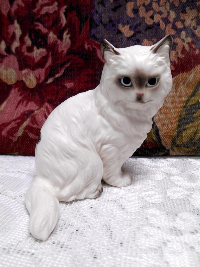 May include: White porcelain figurine of a sitting cat with grey eyes and a fluffy tail.