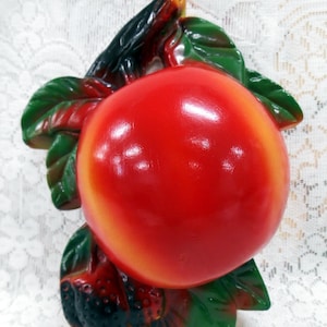 May include: A red and green ceramic peach with leaves. The peach is shiny and has a realistic appearance.