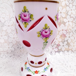 May include: A pink glass goblet with hand-painted floral designs. The goblet features a wide bowl, a stem, and a round base. The bowl is decorated with pink roses and red accents. The base is decorated with red dots and a floral garland.