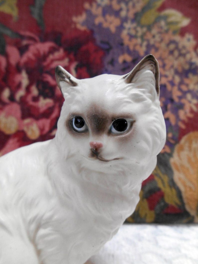 May include: A white porcelain figurine of a cat with brown markings around the eyes and nose. The cat is sitting with its head tilted slightly to the side.