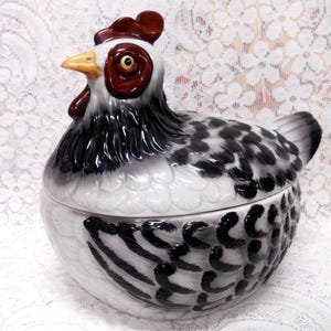 May include: A ceramic hen-shaped container with a lid. The hen is primarily white with black and gray speckled patterns on the body and wings. It has a red comb and wattle, a yellow beak, and brown eyes. The container is likely used for storage or decoration.