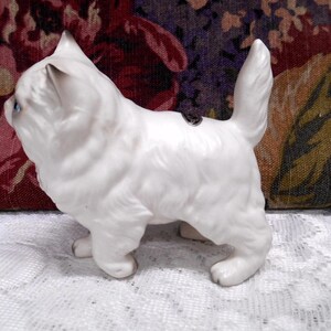 May include: White porcelain figurine of a cat with blue eyes. The cat is standing on all four paws and has its tail curled up.