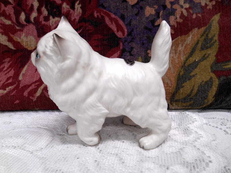 May include: White porcelain figurine of a cat with blue eyes. The cat is standing on all four paws and has its tail curled up.