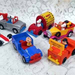 May include: A collection of vintage Peanuts character toy cars. The cars are red, blue, yellow, white, and gray. The cars feature characters like Woodstock, Snoopy, and Charlie Brown.