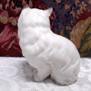 May include: White porcelain figurine of a cat sitting with its head turned to the side. The cat is facing to the left and has a fluffy tail.