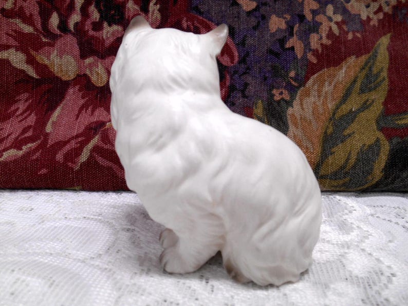May include: White porcelain figurine of a cat sitting with its head turned to the side. The cat is facing to the left and has a fluffy tail.