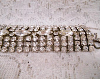 May include: A silver bracelet with multiple rows of small, square-cut clear rhinestones. The bracelet has three larger, oval-shaped rhinestones in the centre. The jewellery is set against a white, textured background.