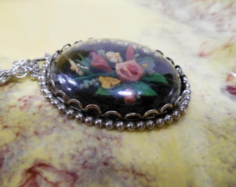 May include: Oval pendant necklace with a silver-tone chain. The pendant features a black background with a floral design in pink, white, and yellow. The pendant is framed with a silver-tone beaded edge.