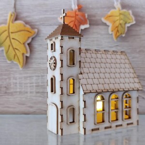 Puede incluir: Una maqueta de iglesia de madera con una cruz y un reloj en la torre. La iglesia tiene ventanas arqueadas y un tejado de tejas. Las ventanas brillan con una luz cálida. Cuelgan recortes decorativos de hojas.