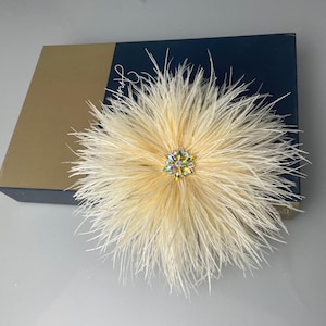 May include: A cream-colored feather flower with a sparkling crystal center. The flower is set on a navy blue and gold box, suggesting a gift or decorative item. The feathers radiate outwards, creating a soft, textured appearance.