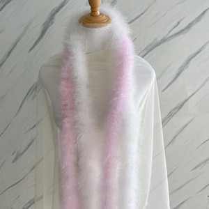 May include: A fluffy, white feather boa with a soft pink stripe down the center. The boa is draped over a white fabric, creating a delicate and elegant look. The boa is a fashion accessory.