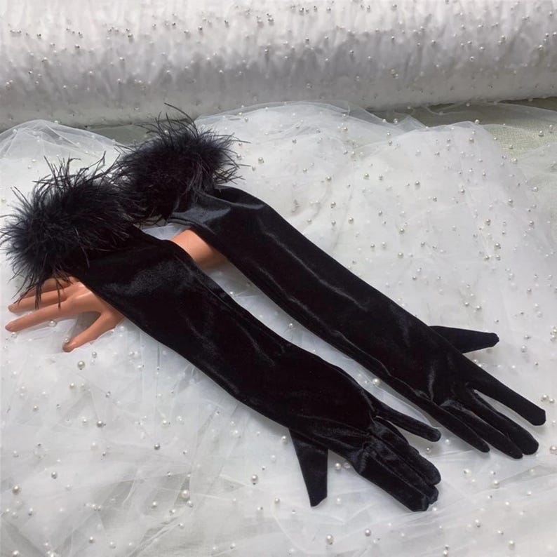May include: A pair of elegant, long black velvet gloves with a feather trim at the wrist. The gloves are displayed on a mannequin hand against a white background.