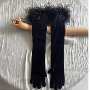 May include: A pair of long, black velvet gloves with a feather trim at the wrist. The gloves are displayed on a wooden hand, showcasing their length and design. The feathers are a dark, fluffy accent.