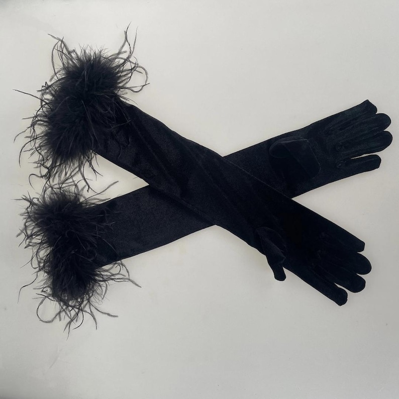 May include: Black opera-length gloves with a soft, velvety texture. The gloves are adorned with fluffy black feathers at the wrist. The gloves are long, extending past the elbow.