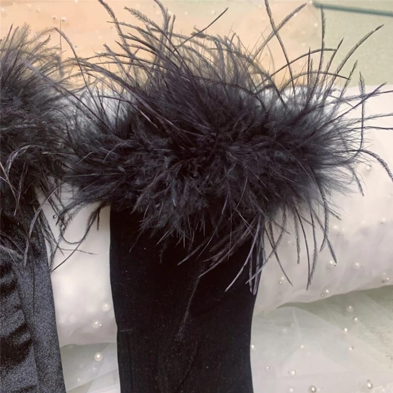 May include: Black feather trim detail on a black garment. The trim is thick and fluffy, with long, wispy feathers. The garment appears to be made of a soft, dark fabric. The background is a soft white fabric.