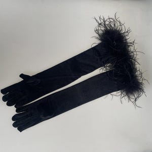 May include: A pair of long, black gloves with a soft, velvety texture. The gloves extend well past the elbow and are trimmed with fluffy black feathers at the top. The gloves are laid out on a white surface.
