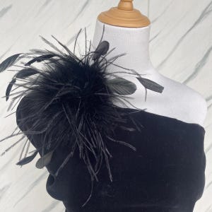 Op de afbeelding: Een zwart veren schouderstuk, waarschijnlijk voor formele kleding, wordt getoond op een witte mannequin. Het accessoire heeft een cluster van pluizige zwarte veren en verschillende langere, strakke zwarte veren die naar buiten uitsteken.