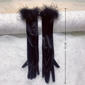 May include: A pair of long, black velvet gloves with a feather trim at the wrist. The gloves measure approximately 23.6 inches (60 cm) in length. The gloves are displayed on a white surface.