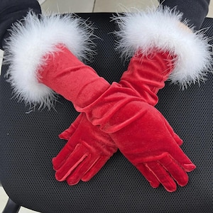 May include: A pair of red velvet gloves with white feather trim at the wrist. The gloves are long, extending past the wrist. The gloves are lying on a black surface.