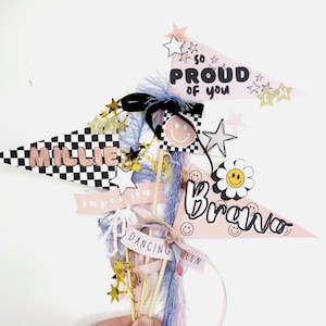 May include: A collection of decorative pennant flags with text and star accents. The flags feature phrases like "Millie", "So Proud of You", and "Bravo". The color palette includes pink, black, white, and gold.