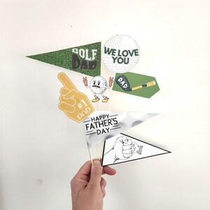 Op de afbeelding: Een verzameling Vaderdagdecoraties met een golfthema. Inclusief een schuimvinger, wimpelvaantjes met tekst en een golfbal met de tekst "We Love You". De decoraties worden door een hand vastgehouden.