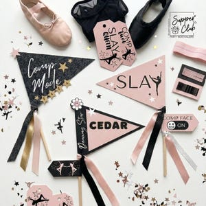 May include: A collection of dance-themed party decorations in pink, black, and gold. Includes pennant flags with text like "Comp Mode" and "Slay", ballet shoes, and hair accessories. Scattered confetti adds a festive touch.