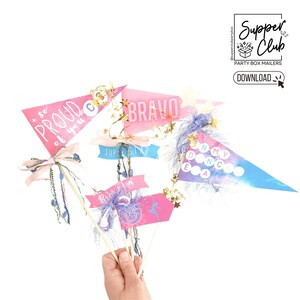 May include: A set of five pink and blue pennant flags with gold glitter and ribbons. The flags have the words "so proud of you", "bravo", "superstar", and "in my dance" printed on them.