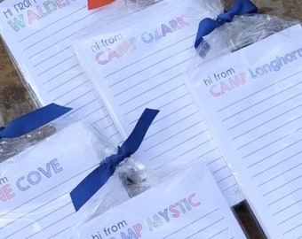 Puede incluir: Cuatro juegos de blocs de notas diferentes envueltos en plástico transparente con cintas azules y rojas. Cada bloc de notas tiene un nombre de campamento diferente impreso en la parte delantera: Camp Waldemar, Camp Ozark, Camp Mystic y Camp Longhorn. Los blocs de notas tienen papel rayado y dicen "Hi from" encima del nombre del campamento.